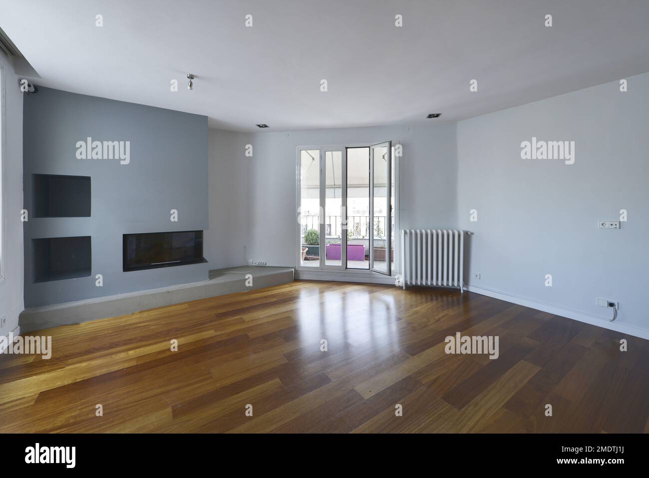 Soggiorno vuoto con pavimento in legno di quercia scuro, pareti lisce blu-grigie, camino a muro e accesso ad una terrazza con tende Foto Stock