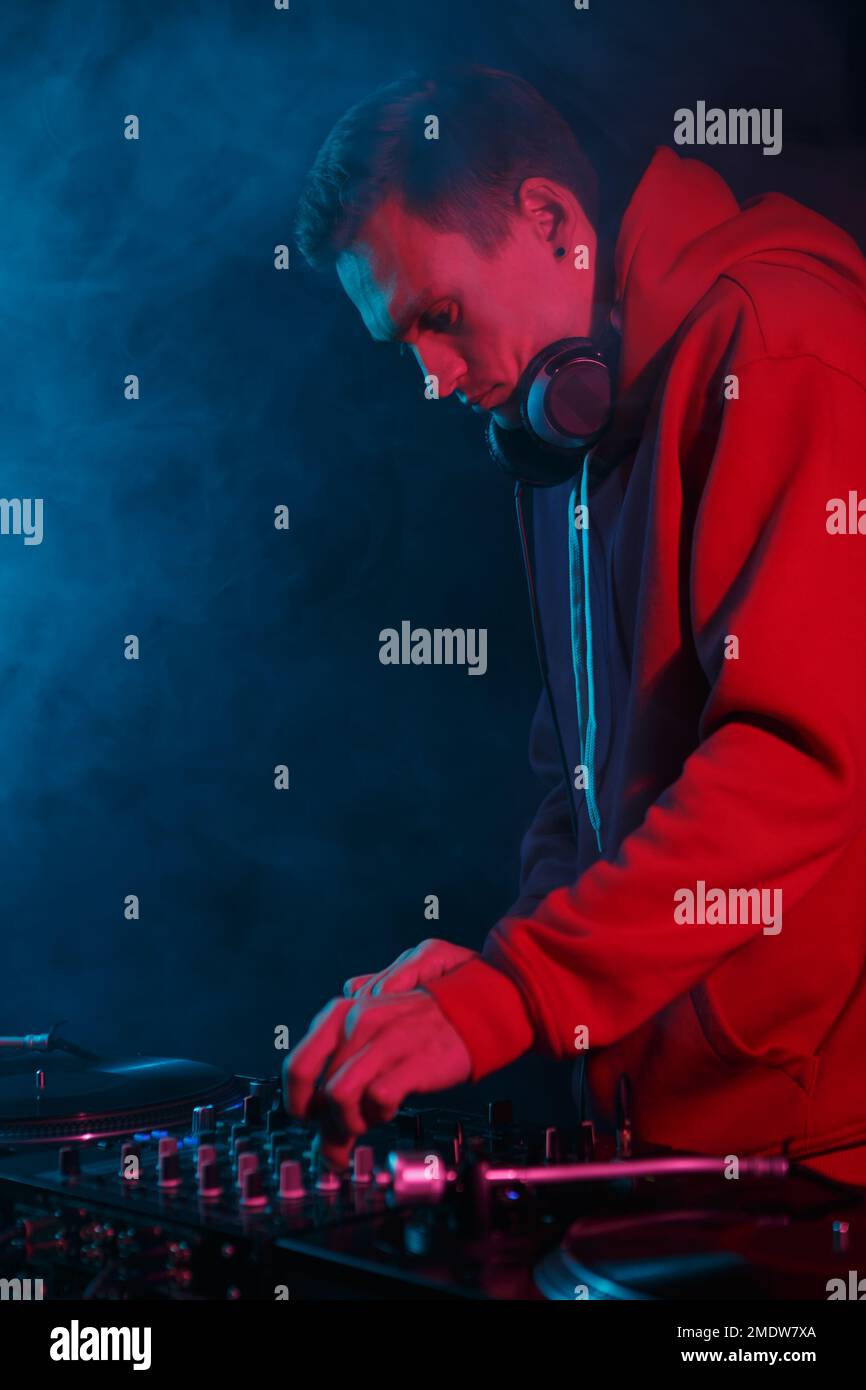 Fresco giovane dj in felpa con cappuccio rosso suona musica durante la festa in fumo. Uomo bianco che gioca nel night club Foto Stock