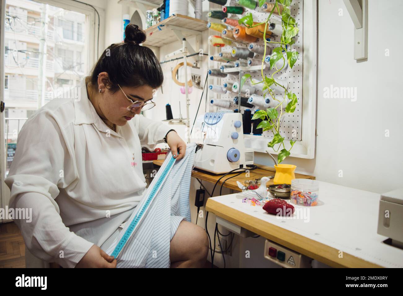 giovane donna di etnia argentina seamstress è seduta nel suo atelier da cucire, lavorando tessuto di misurazione con un metro a nastro, facendo un abito. Foto Stock