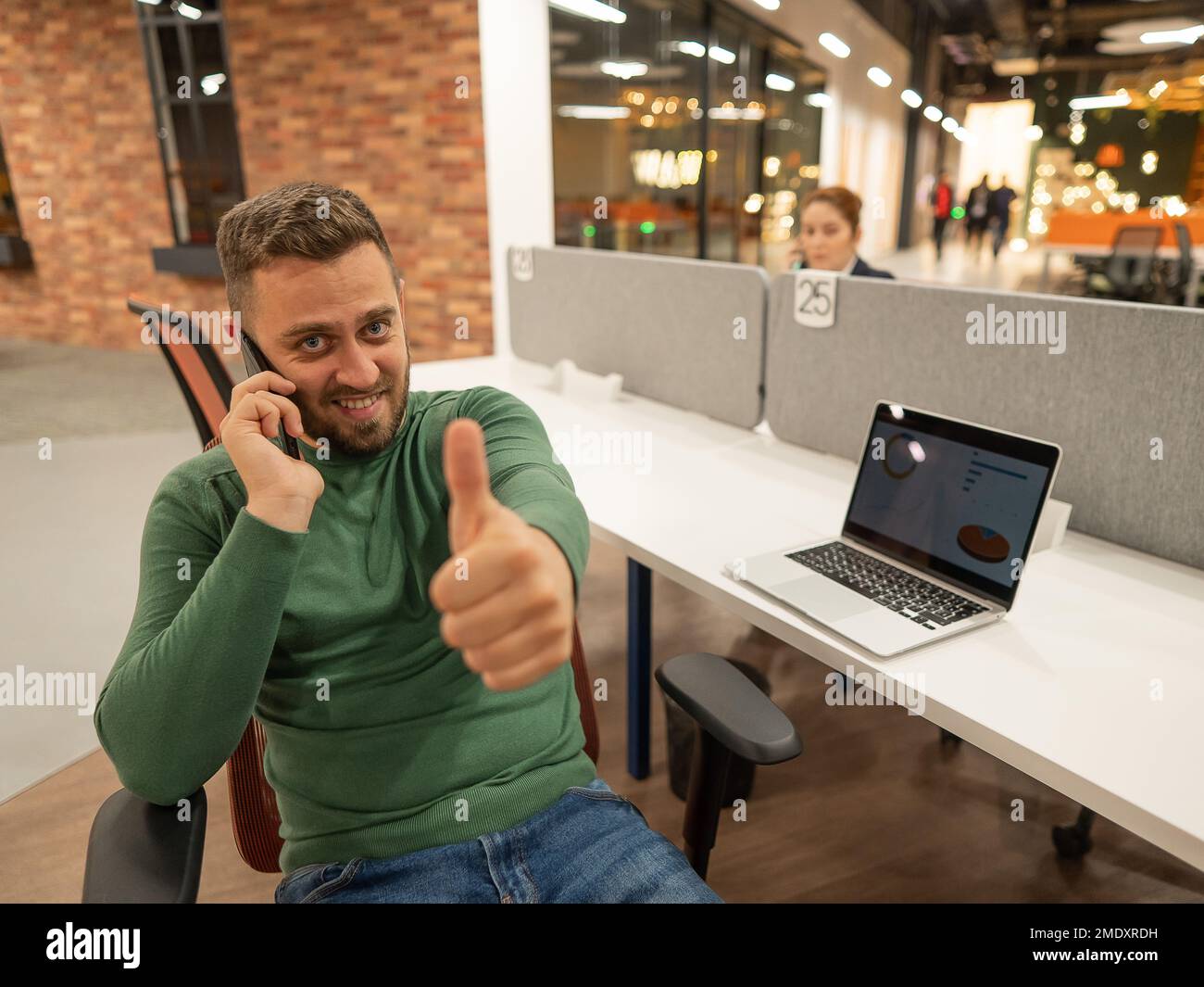 Uomo caucasico che parlava su uno smartphone mentre era seduto alla sua scrivania in un moderno spazio di coworking. Uomo d'affari che guarda la macchina fotografica e mostra i pollici Foto Stock