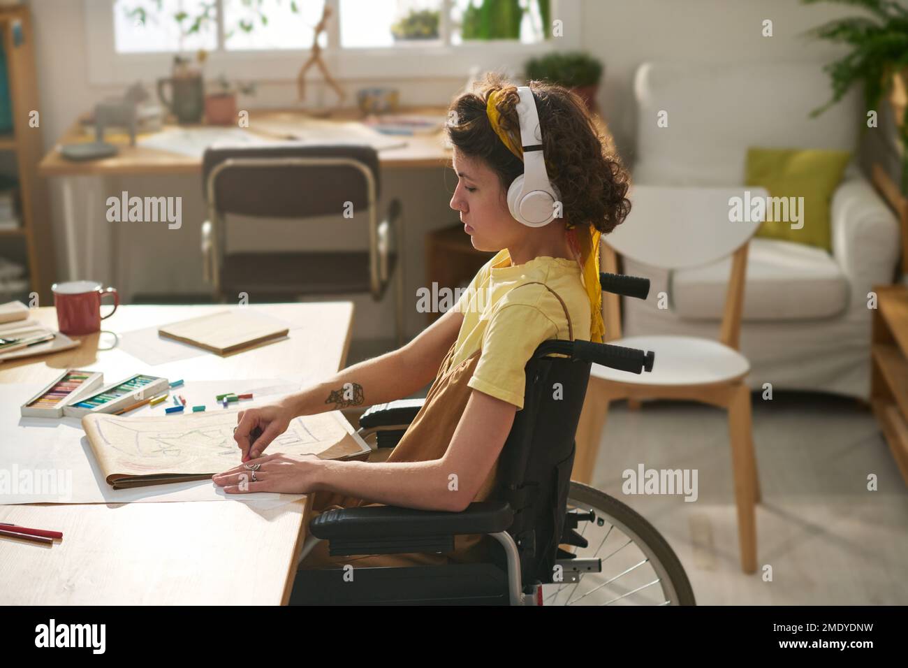 Giovane donna con disabilità che si concentra sulla creazione di nuove opere d'arte mentre si siede in sedia a rotelle e l'ascolto di musica in cuffie Foto Stock