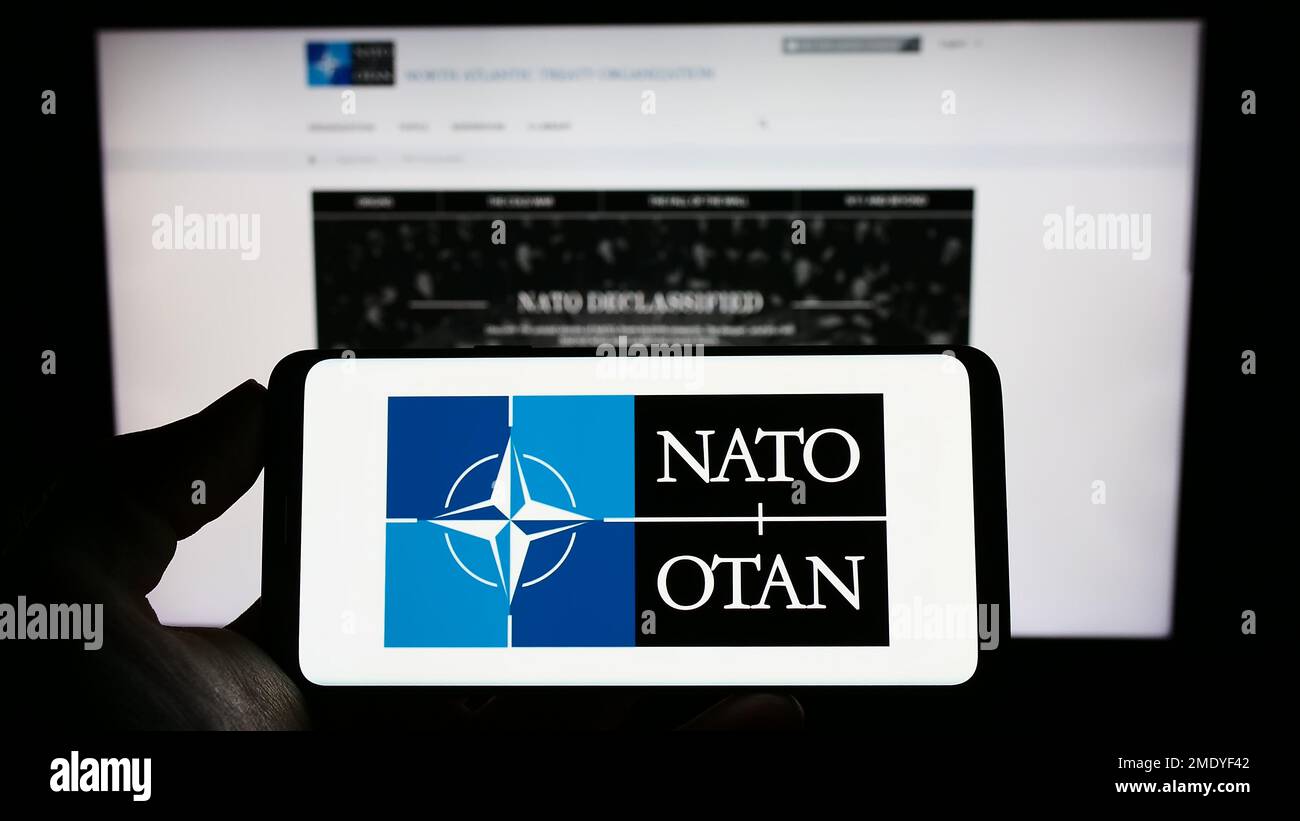 Persona che tiene in mano uno smartphone con il logo della NATO (North Atlantic Treaty Organization) sullo schermo di fronte al sito Web. Messa a fuoco sul display del telefono. Foto Stock