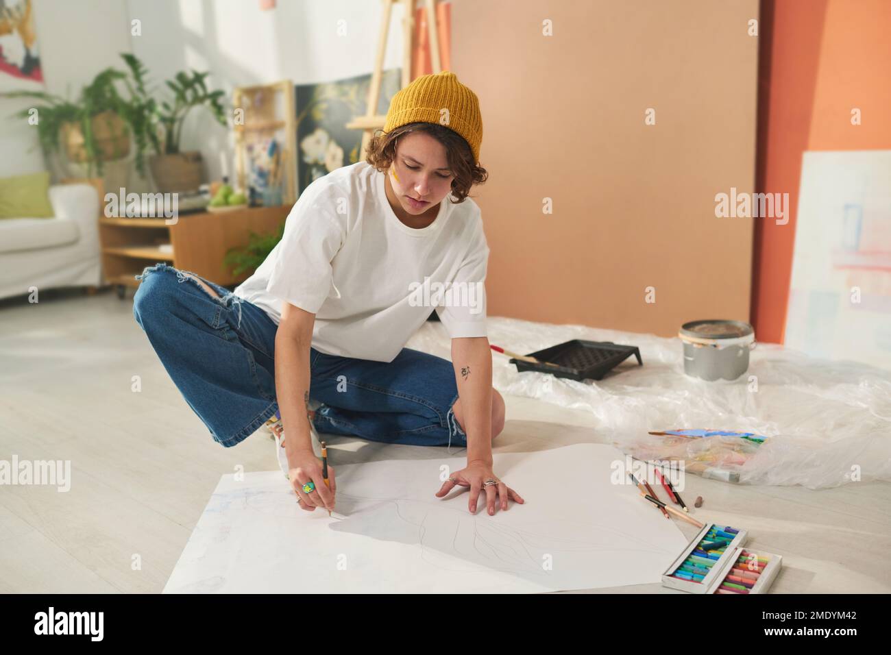 Giovane e creativa bruna artista femminile disegno con pastelli su grande foglio di carta bianca mentre si siede sul pavimento in casa studio Foto Stock