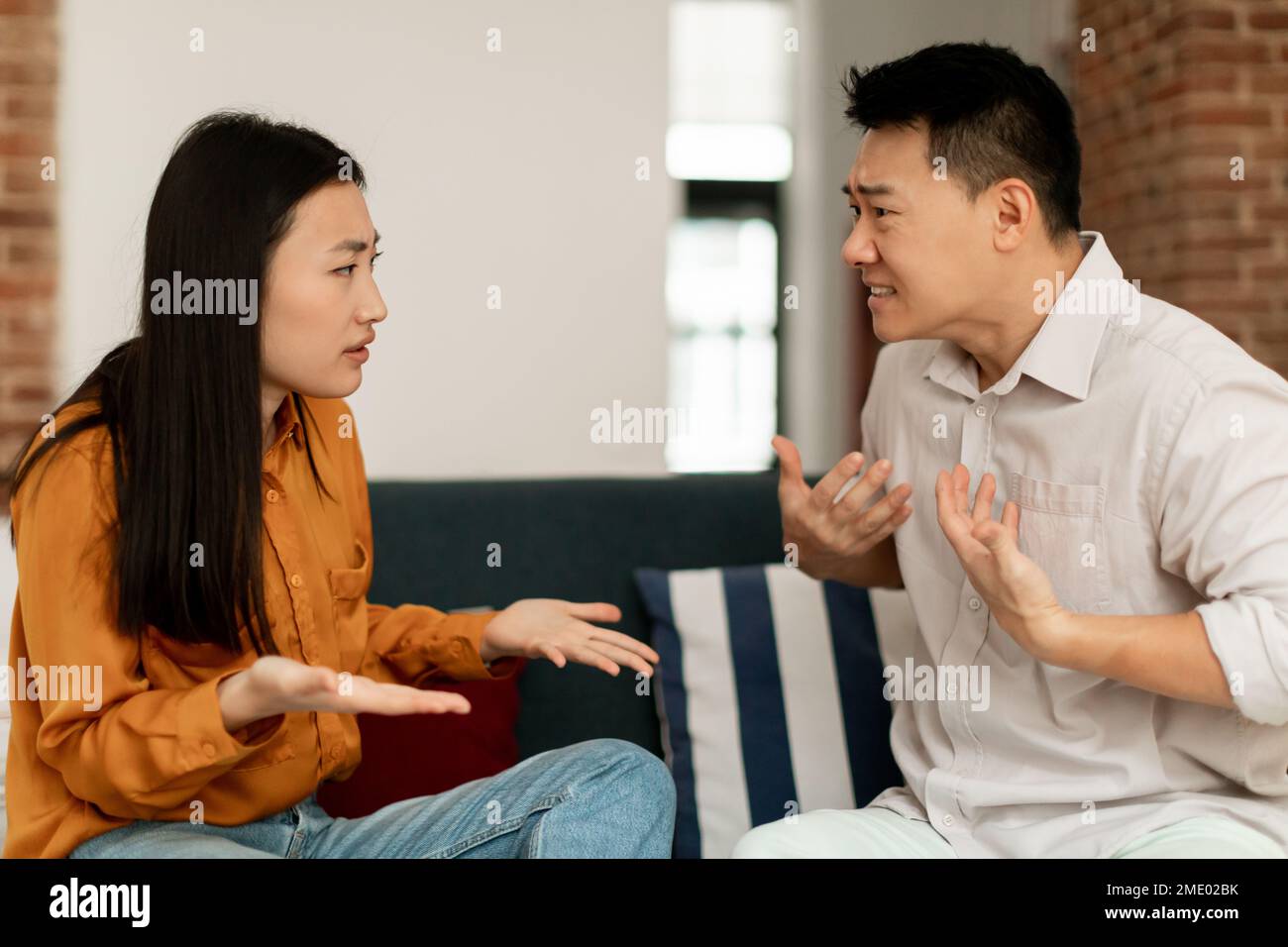 Sposi asiatici arrabbiati che hanno litigato, seduti sul divano e litigando, guardandosi l'un l'altro, vista laterale Foto Stock