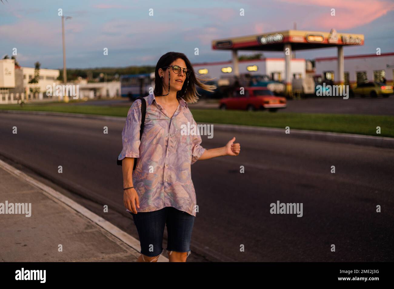 Giovane turista femminile in occhiali con zaino che mostra pollice in su mentre si cattura l'auto su strada al tramonto a Cuba Foto Stock