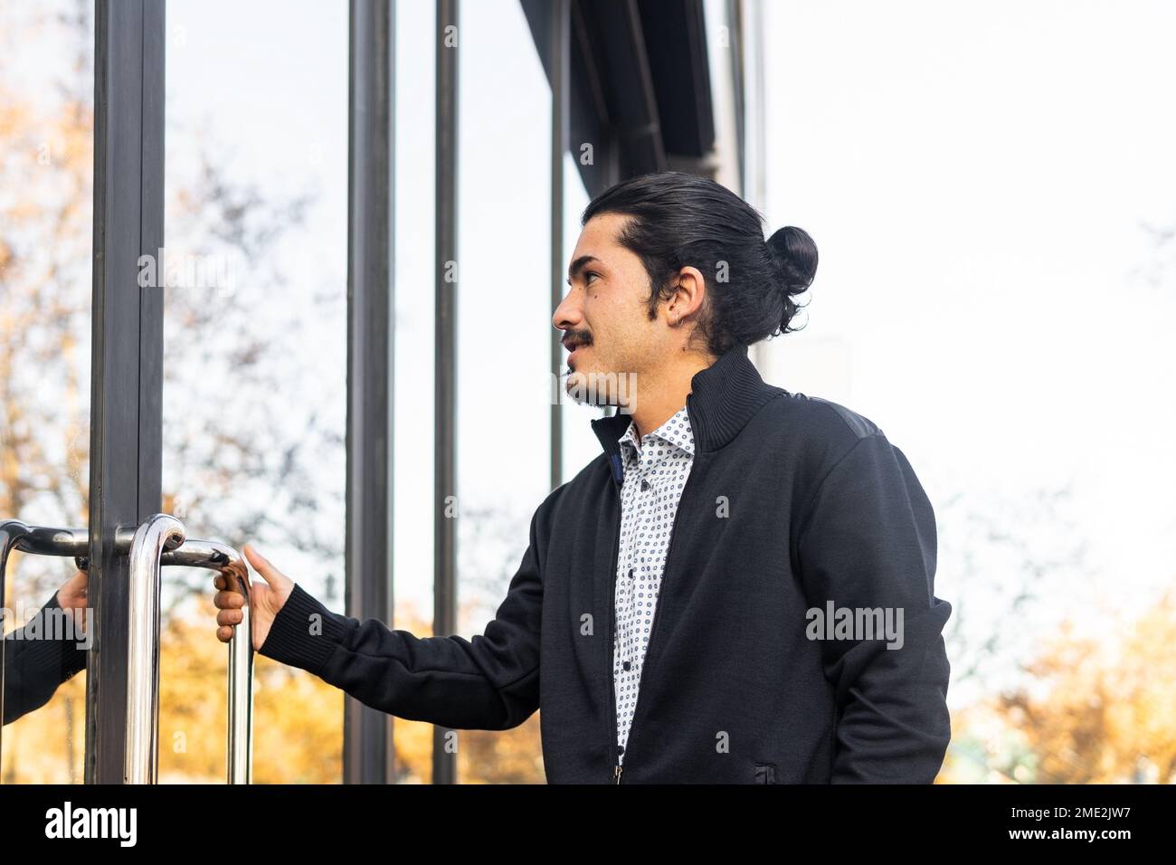 Vista laterale del giovane fidato dipendente maschile ispanico in abiti casual elegante apertura porta di un edificio moderno con pareti in vetro specchiato al sole Foto Stock