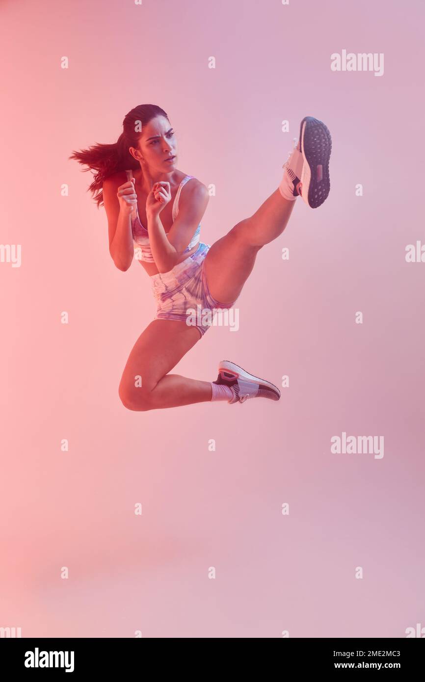 Corpo pieno di determinata giovane combattente femminile con coda di ponytail in colorful sportswear che esegue il calcio di salto su sfondo rosa in studio Foto Stock
