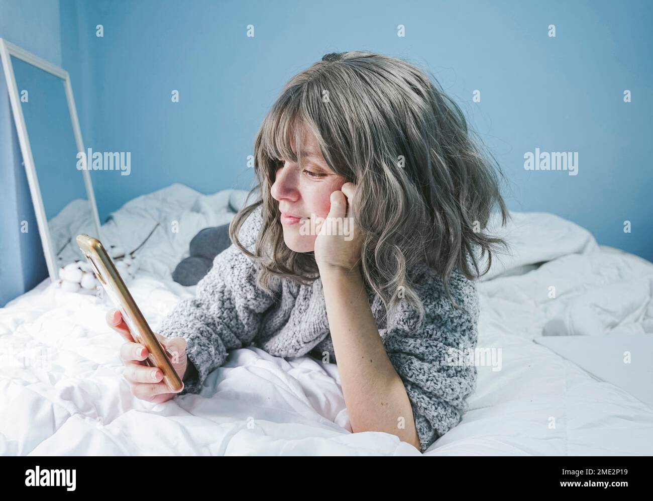 Donna adulta positiva con capelli ricci corti grigi in caldo maglione sdraiato su comodo letto e smartphone browser in camera da letto con specchio e parete blu Foto Stock