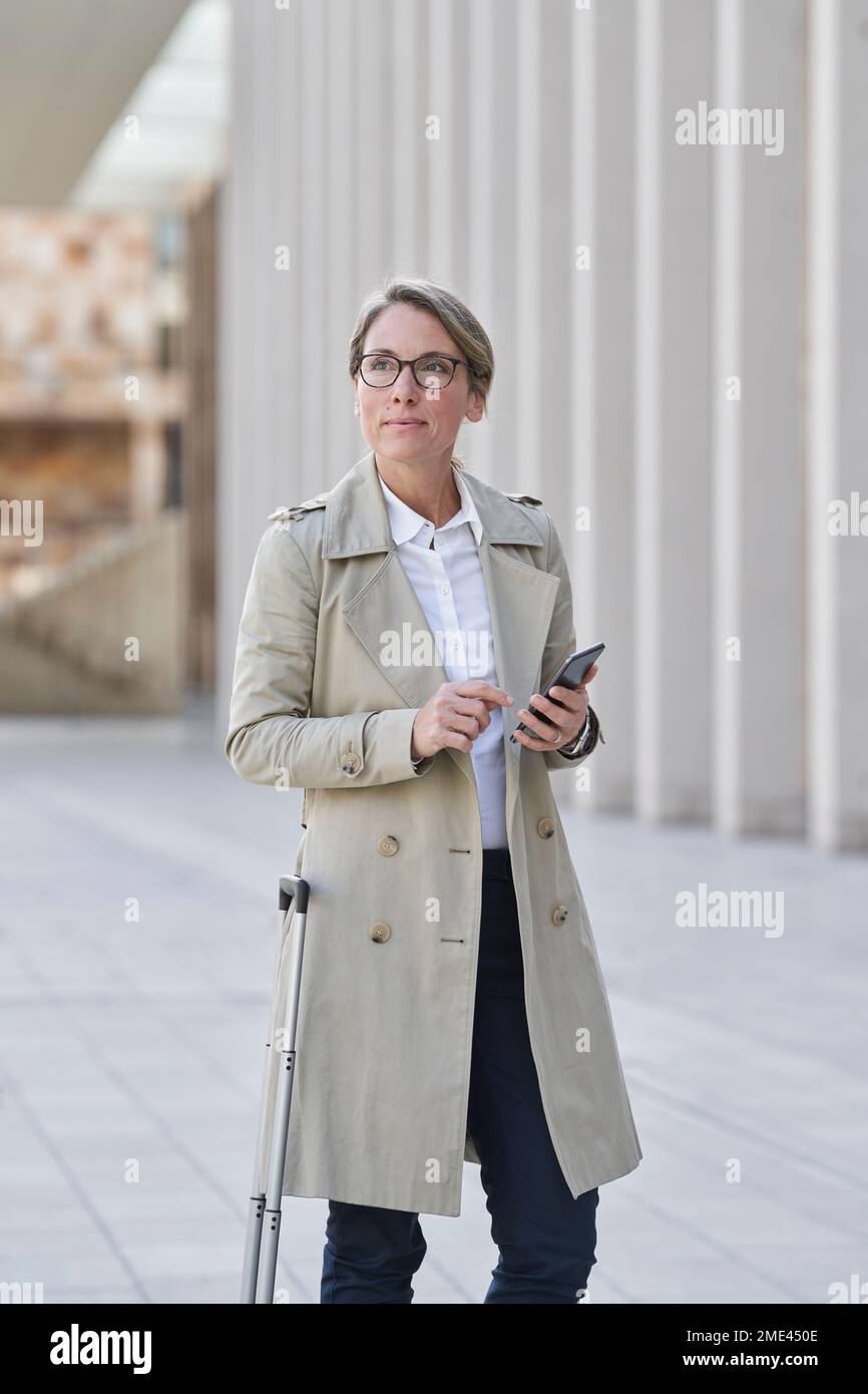 Donna d'affari matura con bagaglio a rotelle in piedi in città con smartphone Foto Stock
