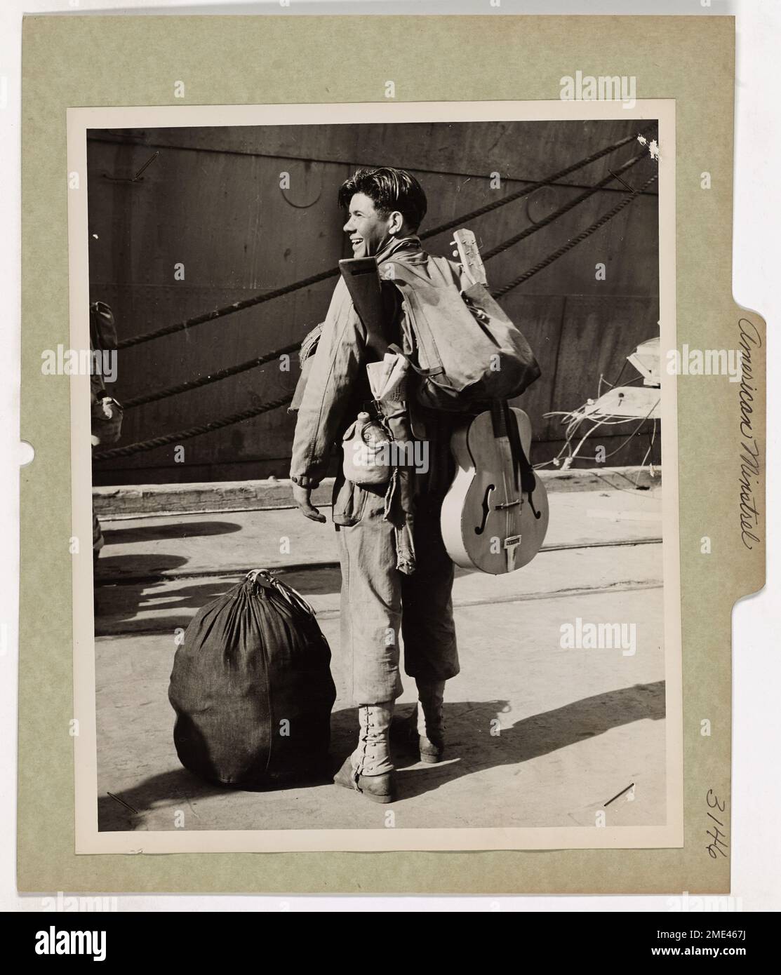 Un soldato americano, che si prepara a salire a bordo di un trasporto di combattimento con equipaggio della Guardia Costiera nel Pacifico meridionale, porta il suo fucile e la sua chitarra. La sua prontezza alla battaglia è evidente quando indossa un sorriso, simboleggiando sia le difficoltà che lo spirito dei soldati durante la guerra. Foto Stock