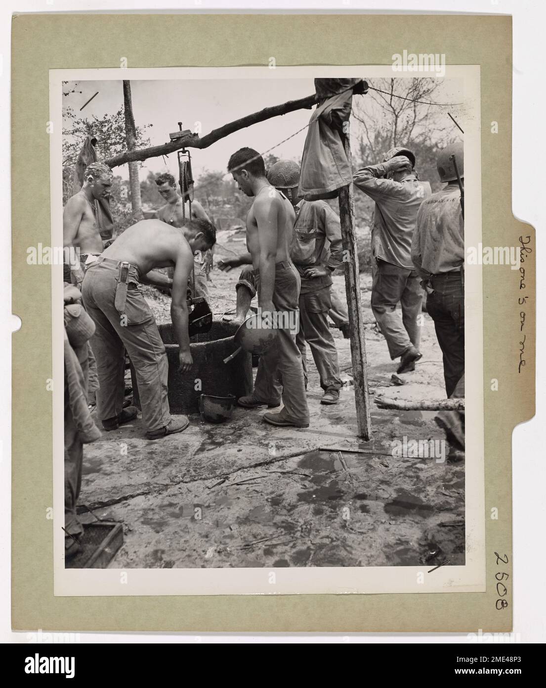 In un pozzo giapponese sull'isola di Saipan, le guardie costiere e i soldati statunitensi condividono un drink dai loro caschi da battaglia, che servono anche come lavabi. La scena informale riflette la cameratismo e l'intraprendenza delle forze americane mentre navigavano nella vita quotidiana durante la campagna di salto delle isole nel Pacifico. Foto Stock