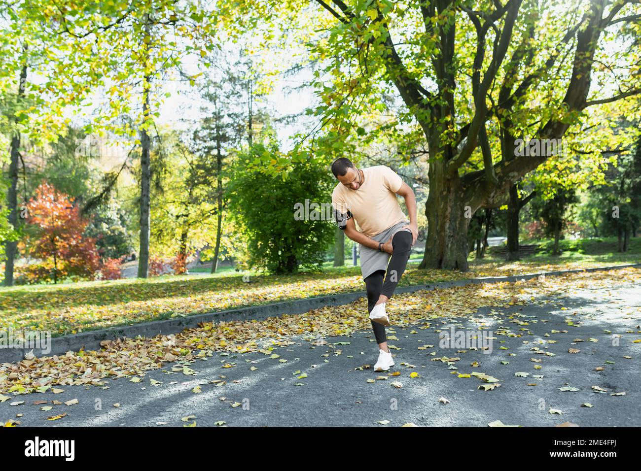 Runner ferito la gamba mentre jogging nel parco vicino agli alberi, l'uomo ispanico ha spasmo muscolare massaggiando il ginocchio, l'uomo è in esecuzione e l'esercizio di fitness in una giornata di sole. Foto Stock