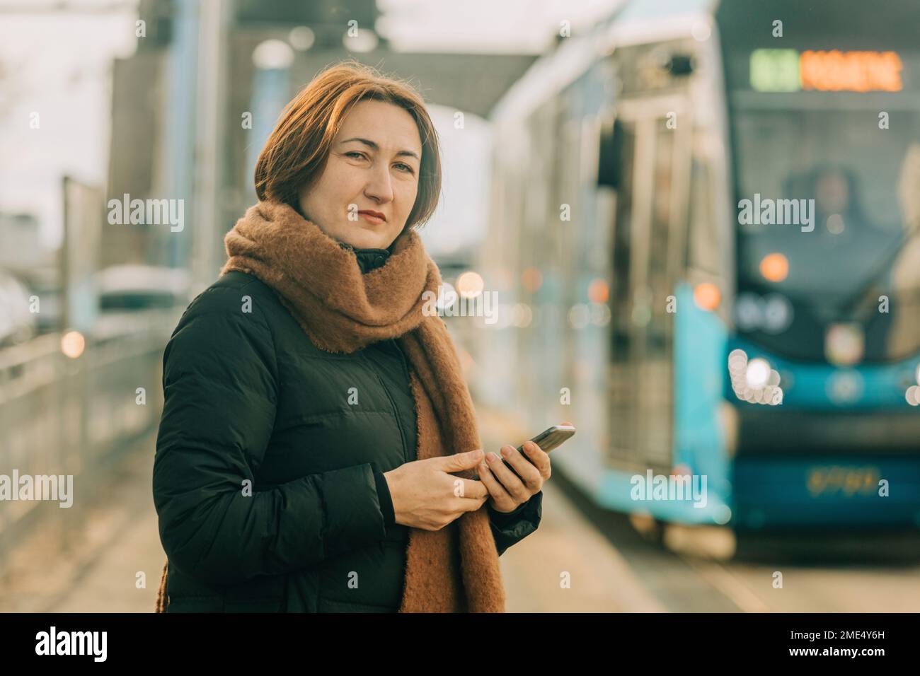 Donna matura sorridente in piedi con smartphone alla stazione del tram Foto Stock
