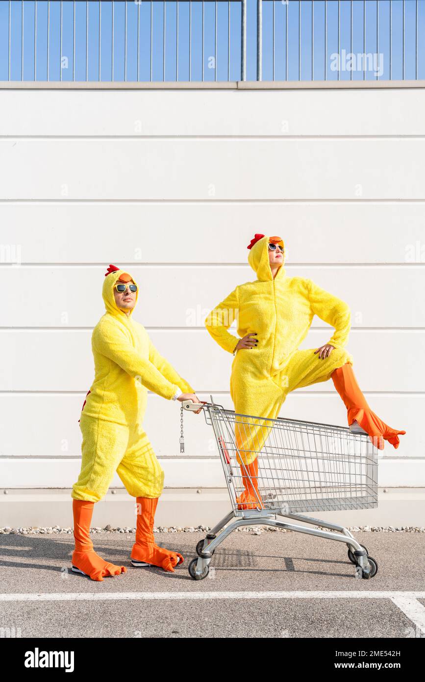 Amici che indossano costumi di pollo in piedi con carrello per la spesa di fronte al muro Foto Stock