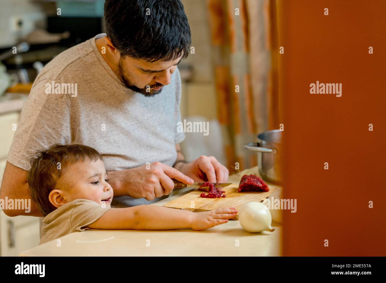Uomo che taglia la carne per figlio raggiungendo la cipolla bianca sul bancone della cucina Foto Stock