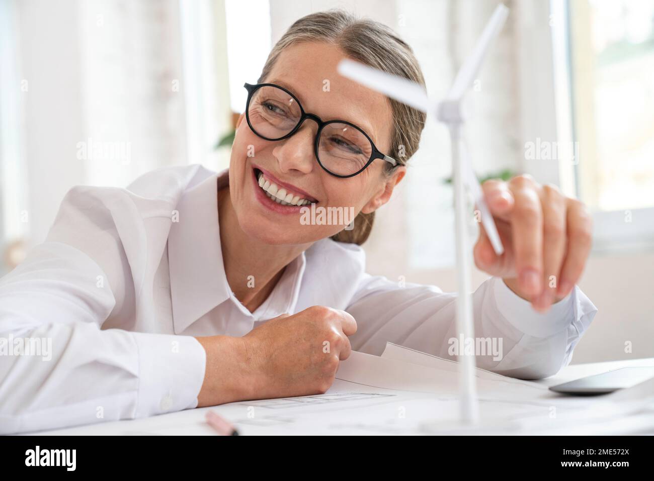 Felice donna d'affari matura che tocca turbina eolica in ufficio Foto Stock