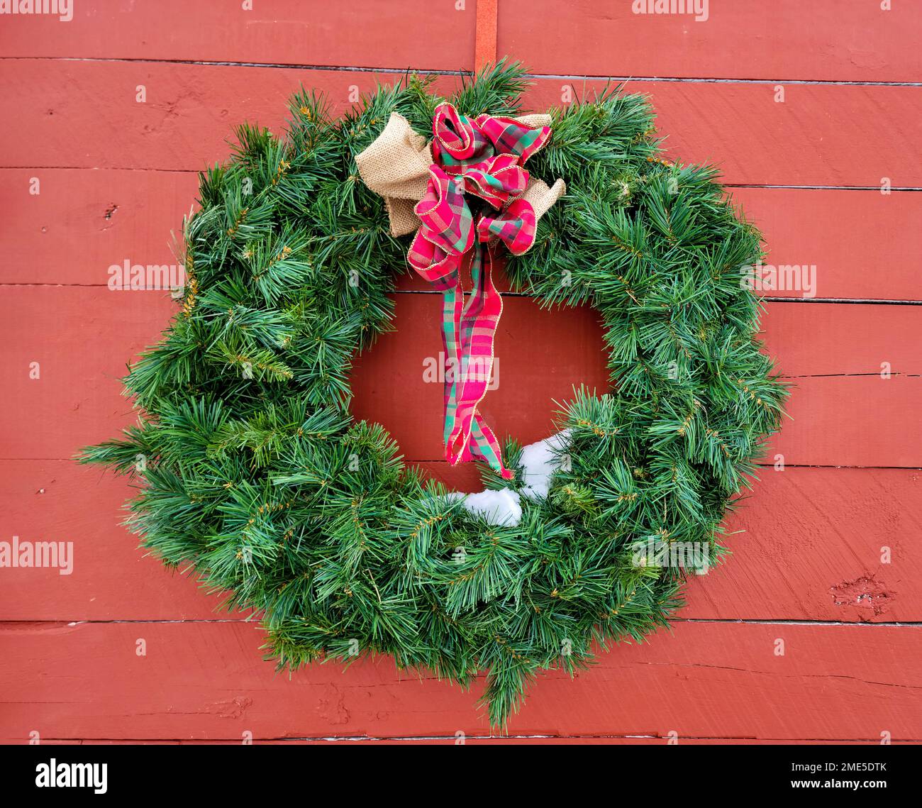 Verde corona di Natale con un arco a plaid e neve sciogliente su un fienile rosso Foto Stock