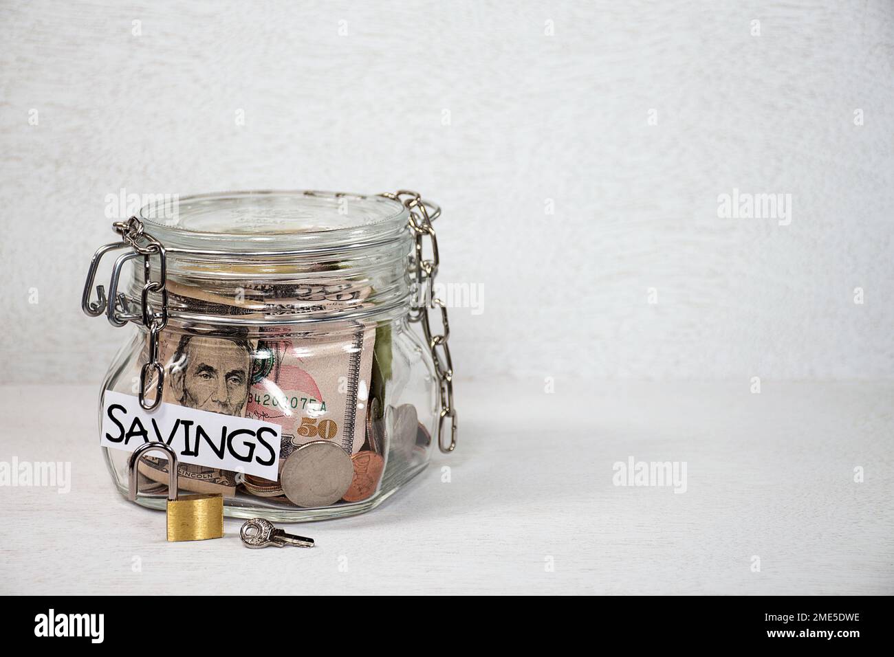 Catena allentata su un vaso di vetro con un'etichetta di risparmio, denaro, lucchetto aperto e chiave su sfondo grigio chiaro testurizzato Foto Stock