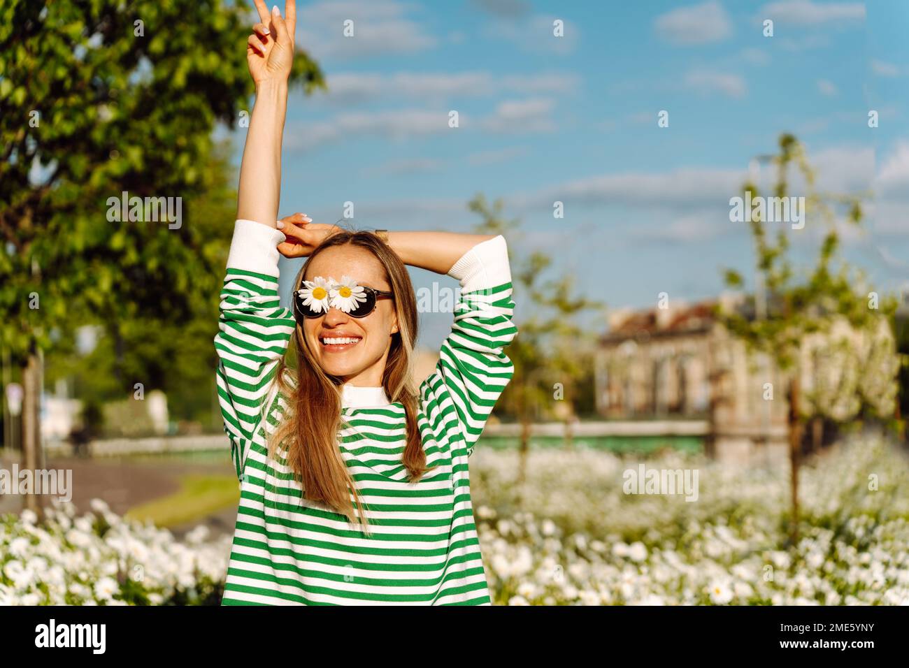 Una donna dai capelli lunghi con margherite sugli occhiali sorride felicemente con la mano in su. Foto Stock