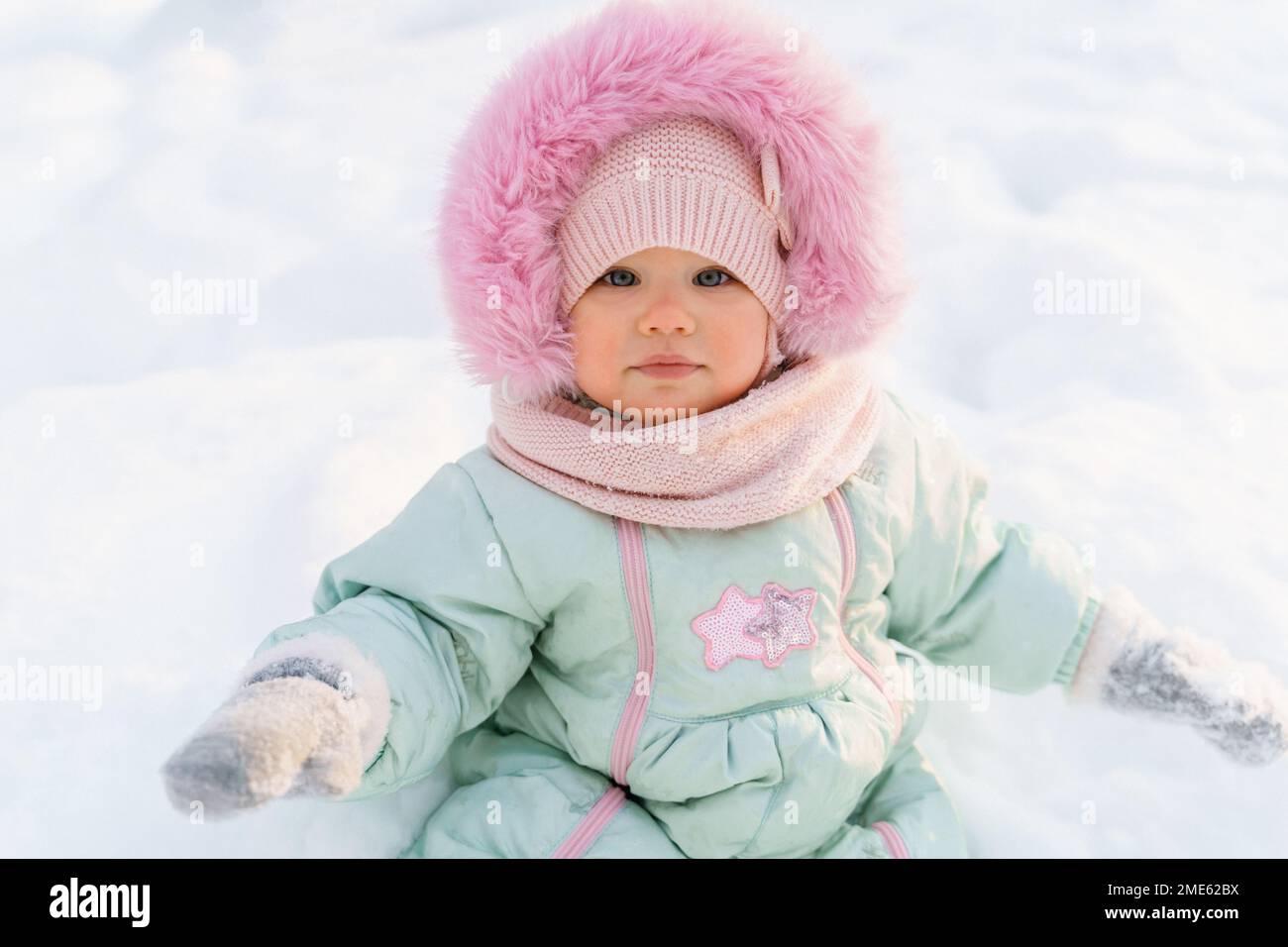 Primo piano ritratto di una ragazza carina e ruddy bambino bambino in un caldo mint-colorato downy complessivo con pelliccia rosa si siede sulla neve bianca in bel tempo soleggiato gelido. Foto Stock