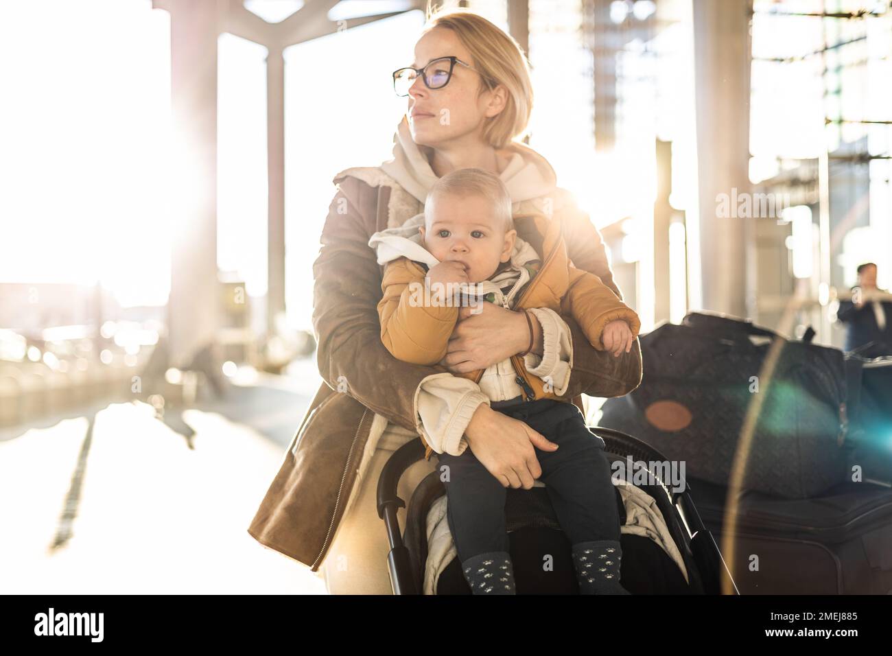 Motherat viaggia con il bambino, camminando, spingendo passeggino e carretto bagagli davanti alla stazione del terminal aeroportuale Foto Stock