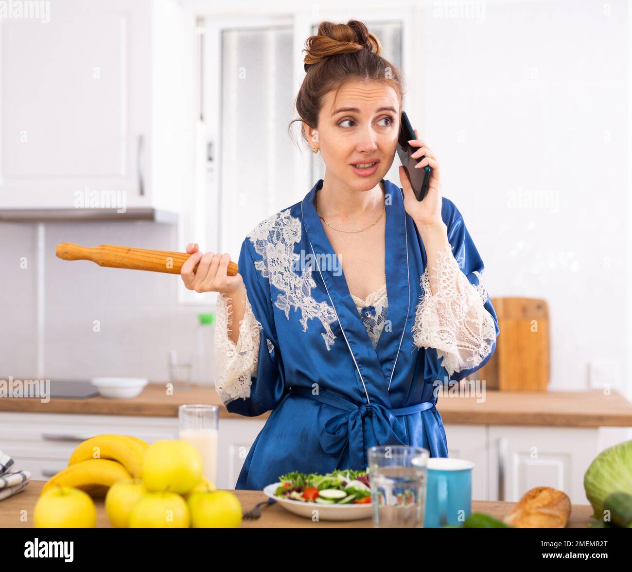 Casalinga insoddisfatta con un perno di rotolamento nelle sue mani che parla sul telefono cellulare in cucina Foto Stock