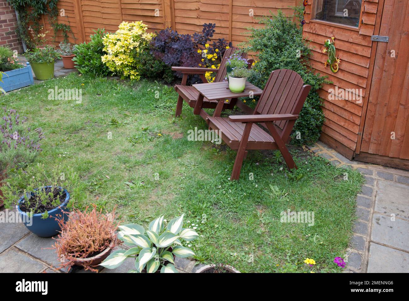Piccolo giardino con capannone, prato turfed e pavimentazione circostante, con sedie da giardino, un piccolo tavolo, e una varietà di piante che crescono in pentole Foto Stock