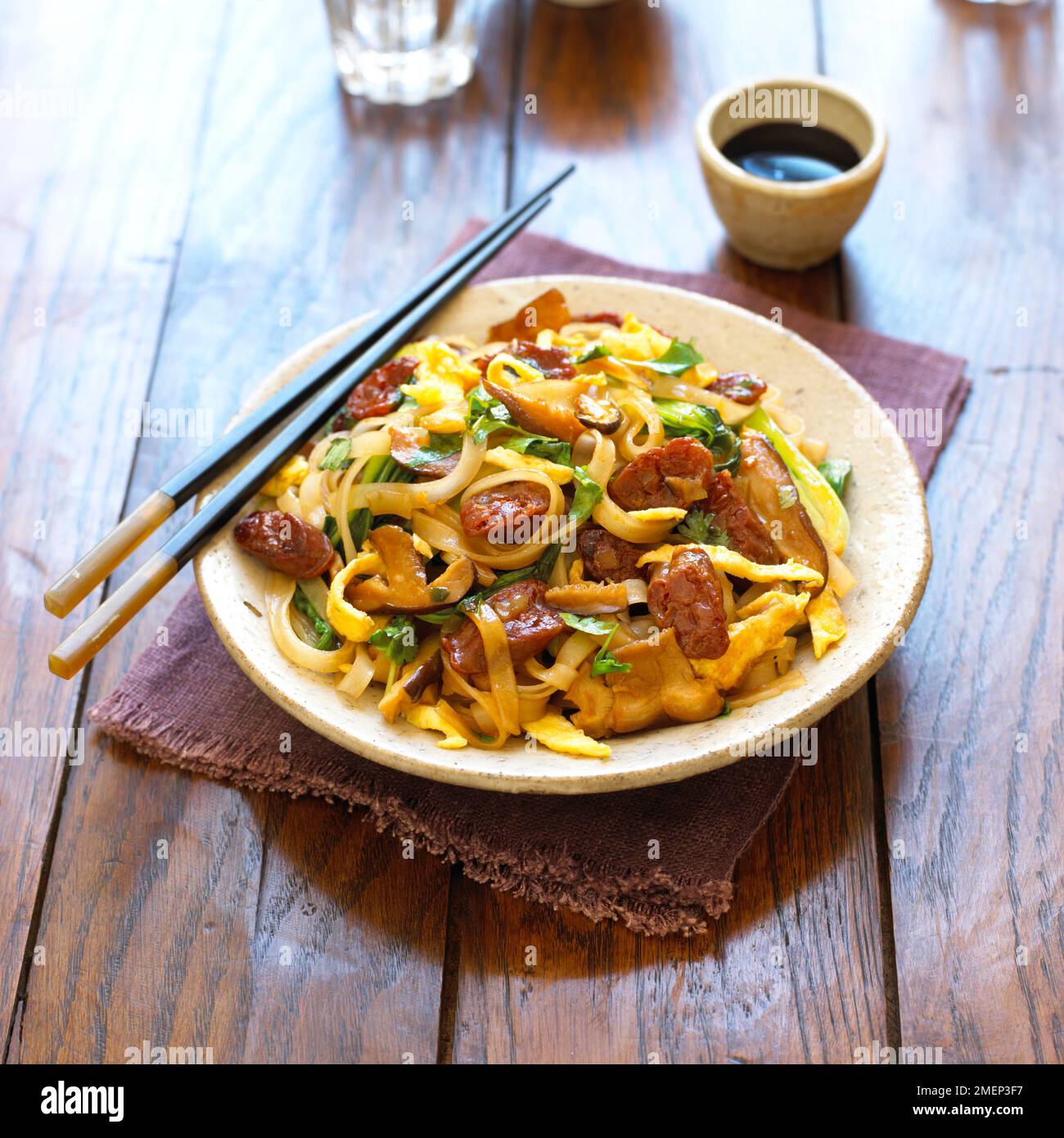 Spaghetti fritti con salsiccia cinese e shiitake, serviti in un recipiente ampio con bastoncini e una ciotola di salsa di soia Foto Stock
