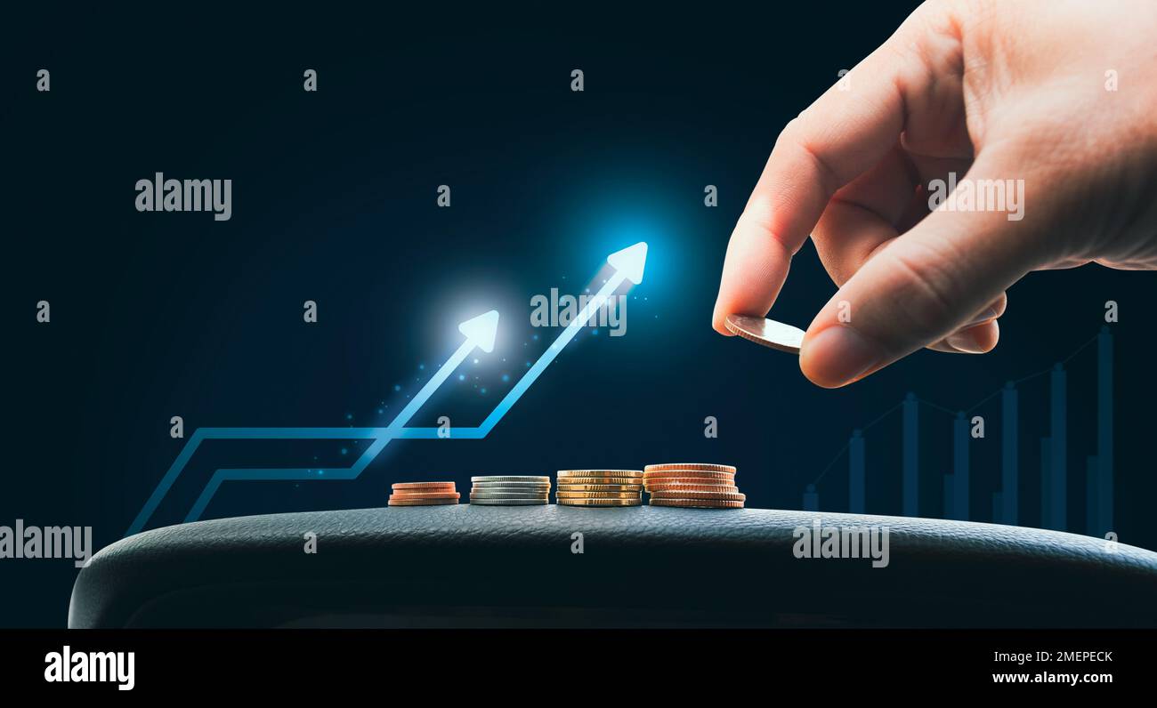 La mano sta cadendo la moneta sulla pila delle monete con la freccia crescente e la tabella di affari sfocata sullo sfondo, l'investimento ed il concetto di risparmio dei soldi Foto Stock