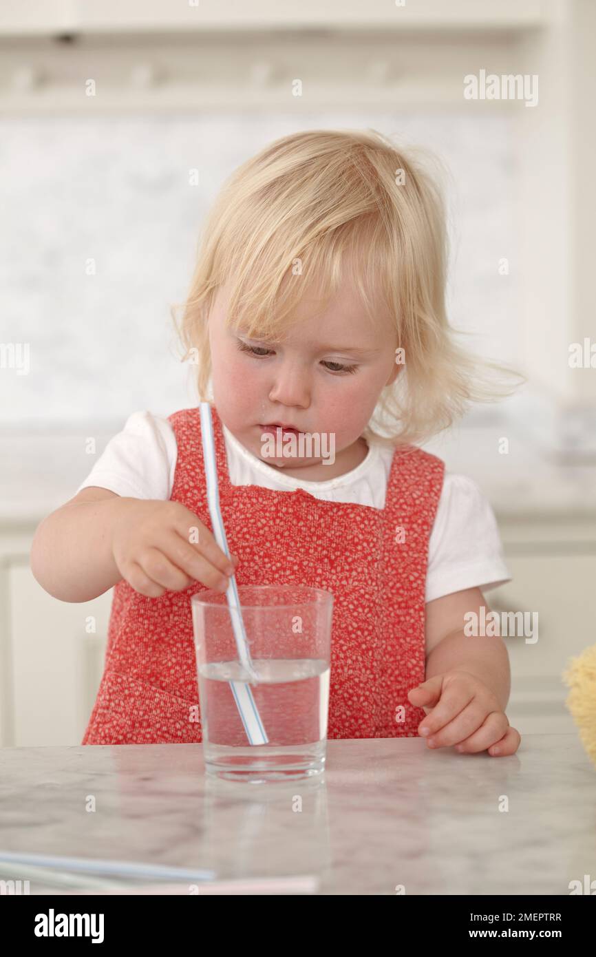 Giovane ragazza che tiene paglia in un bicchiere d'acqua, 18 mesi Foto Stock