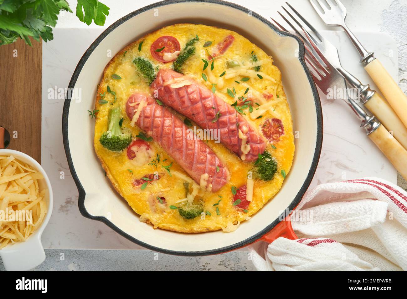 Omelette o frittata con due salsicce, formaggio fuso, pomodori ciliegini, broccoli e prezzemolo su padella rossa o teglia di ferro su fondo bianco di cemento vecchio Foto Stock