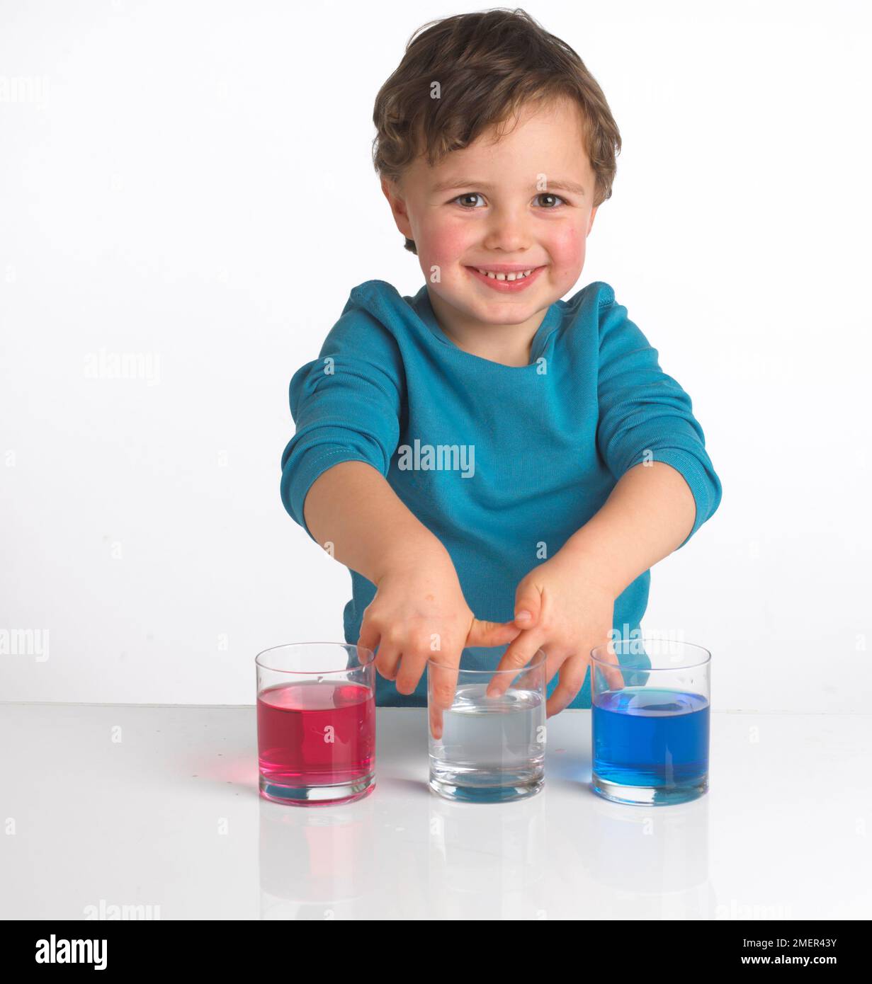 Ragazzo che indossa la parte superiore blu davanti a tre bicchieri contenenti liquido rosso, liquido trasparente e liquido blu, 4 anni Foto Stock
