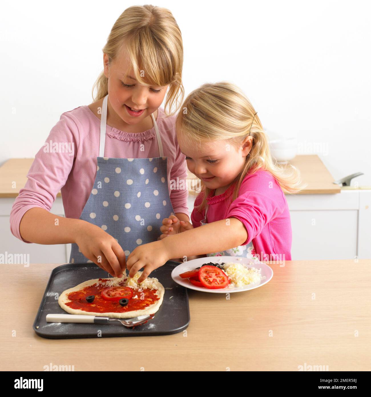Due ragazze mettendo crema sulla pizza cruda base, 7 e 3 anni Foto Stock
