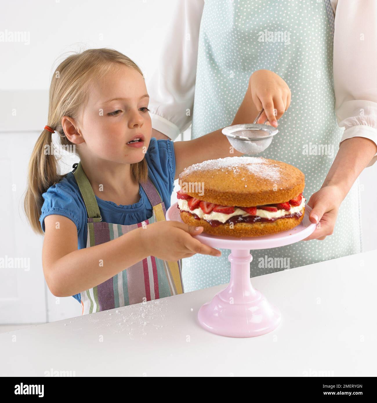 Ragazza setacciando lo zucchero a velo su una spugna di fragola, 5 anni Foto Stock