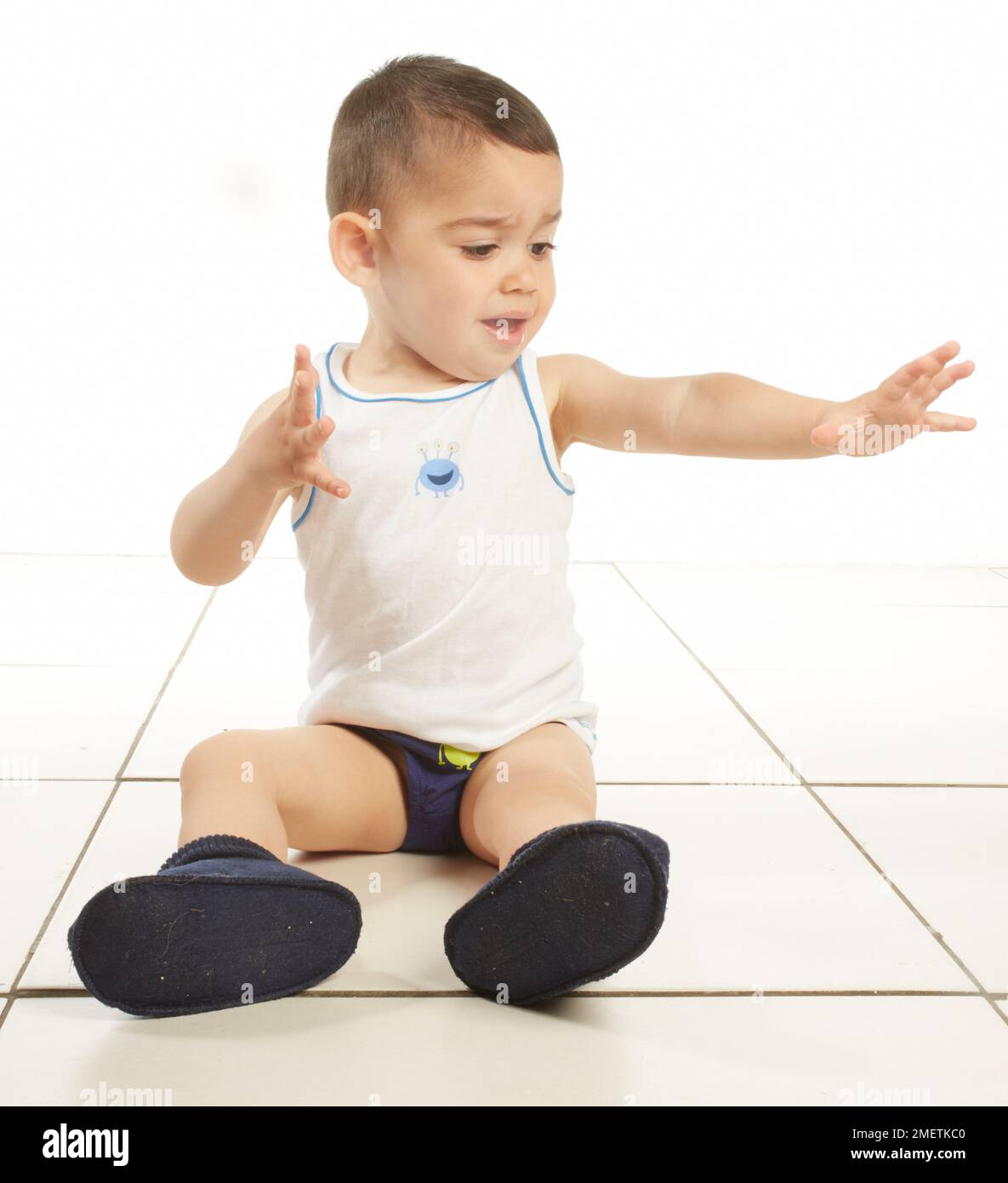 Ragazzo che indossa giubbotto, pantaloni e ciabatte, seduto sul pavimento, 15 mesi Foto Stock