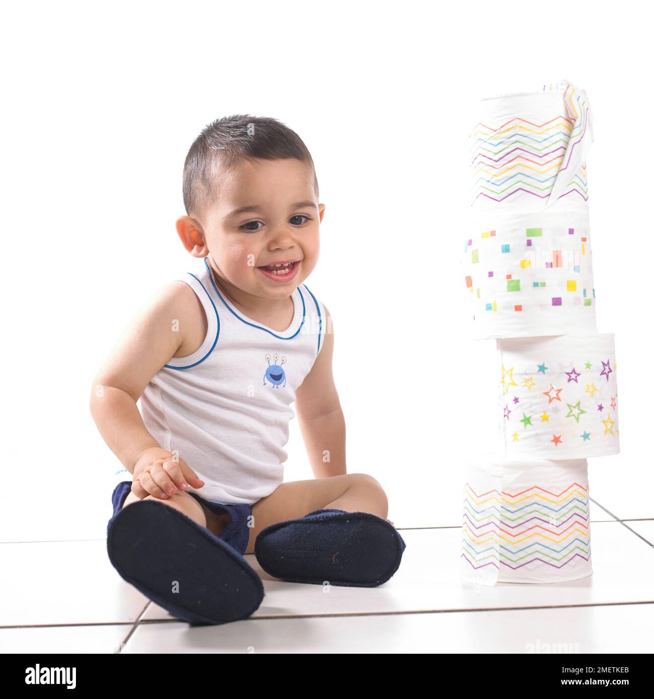 Ragazzo che indossa giubbotto, pantaloni e ciabatte accanto alla torre di rotoli di carta igienica, 15 mesi Foto Stock