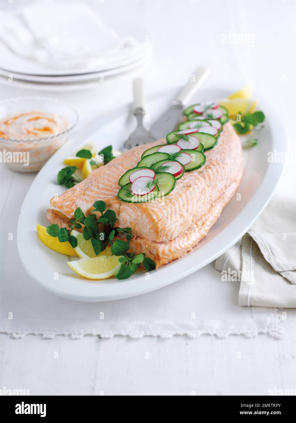 Salmone intero in camicia guarnito con fette di cetriolo, ravanello, crescione e spicchi di limone, servito con maionese al peperoncino Foto Stock