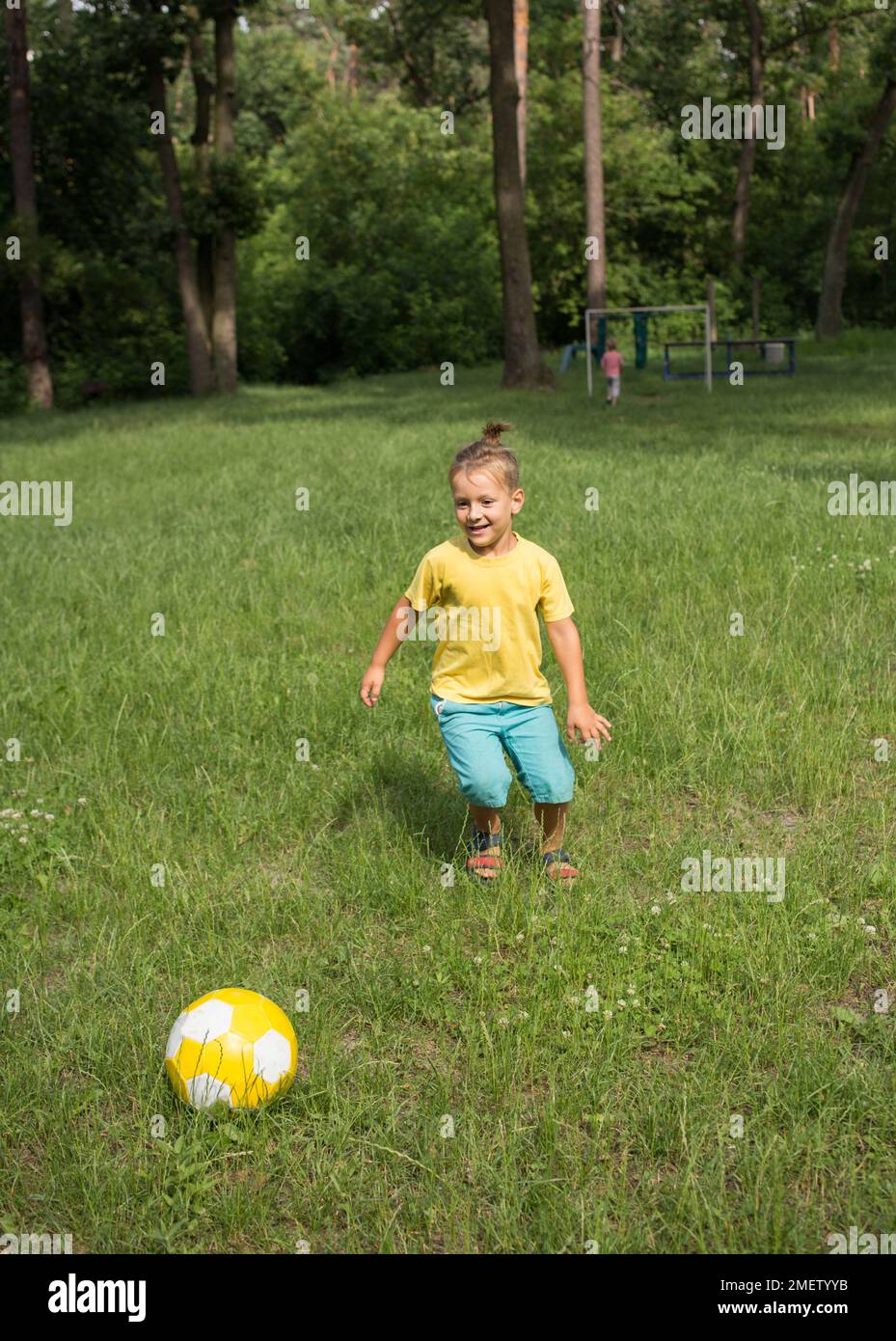 ragazzo di 5-6 anni corre nel parco su erba verde e calcia una palla di calcio gialla. Stile di vita attivo, attività divertenti per i bambini nell'outdo estivo Foto Stock