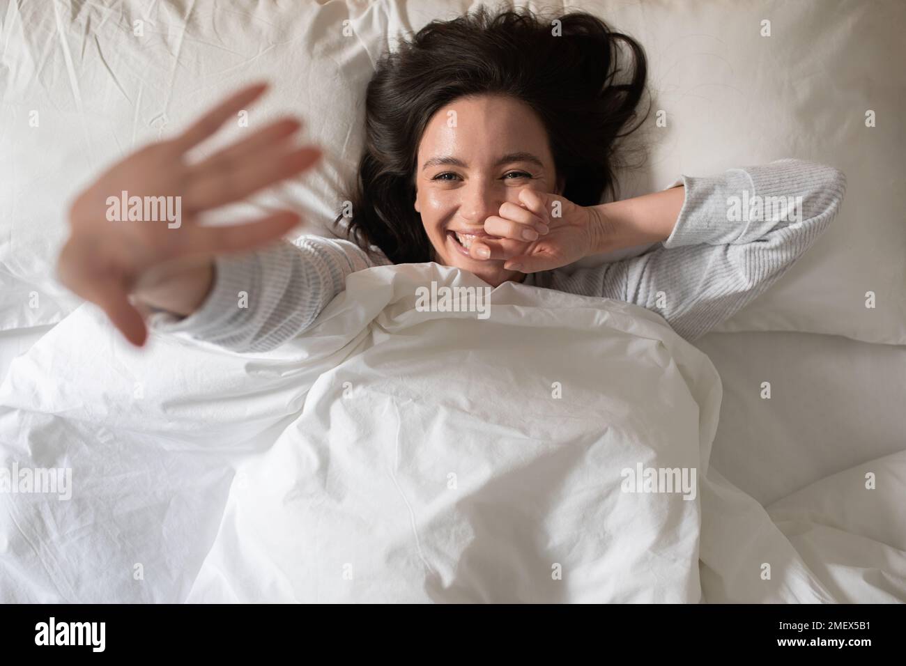 Allegra bella giovane donna europea si sveglia, ondeggia la mano o si chiude dalla macchina fotografica, si trova su un letto bianco Foto Stock