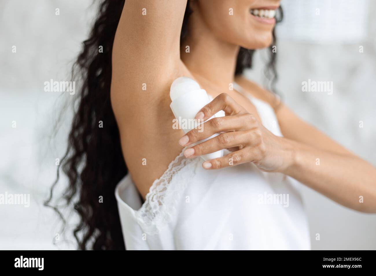 Allegra donna araba millenaria con capelli ricci lunghi in abiti domestici applicando deodorante sulle ascelle Foto Stock