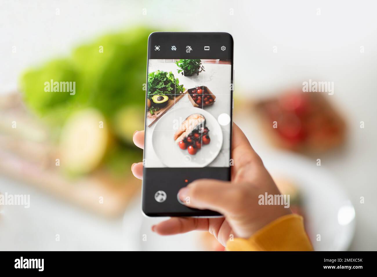 Donna afroamericana che fa foto di piatto con pesce sullo schermo dello smartphone sul tavolo con verdure fresche Foto Stock