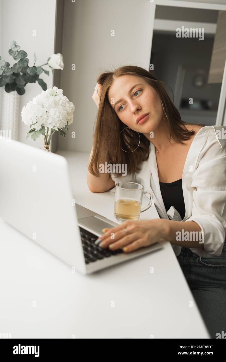 Primo piano di una giovane bella donna che beve il tè mentre si fa una pausa dopo aver digitato e navigato online sul computer portatile mentre si è seduti in una stanza accogliente Foto Stock