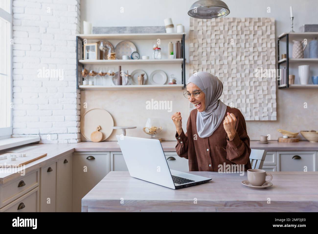 Freelancer in hijab lavorando sul laptop in remoto da casa, donna ha ricevuto buone notizie e buone notizie on-line notifica, felice e celebrando i buoni risultati di realizzazione, donna d'affari in casa ufficio. Foto Stock