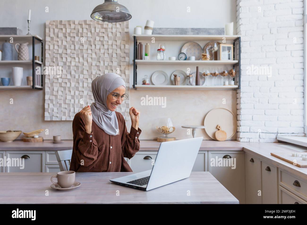 Freelancer in hijab lavorando sul laptop in remoto da casa, donna ha ricevuto buone notizie e buone notizie on-line notifica, felice e celebrando i buoni risultati di realizzazione, donna d'affari in casa ufficio. Foto Stock