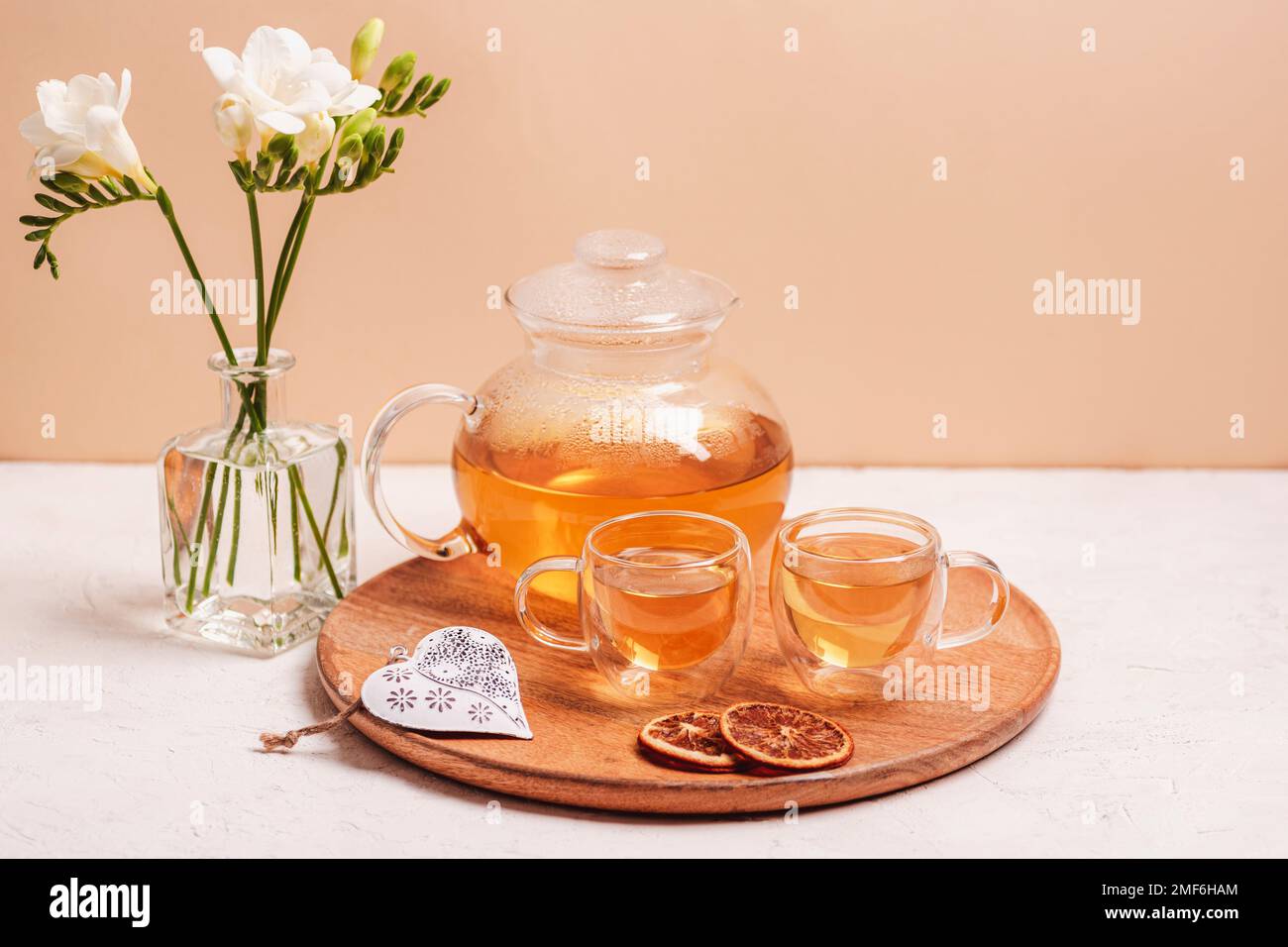 Tè verde in teiera di vetro e tazze sul tavolo. Teiera e tazze di tè su vassoio di legno, cuore giocattolo, fette di arancia e fiori di freesia in vaso. Spazio di copia. Foto Stock