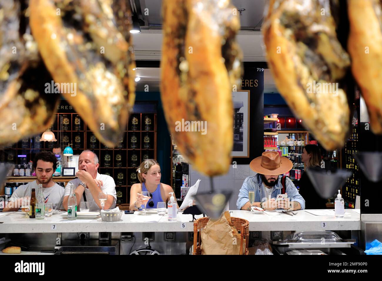 I clienti pranzano in un chiosco di cibo spagnolo con prosciutti iberici e Jamon appesi in primo piano.le Gourmet. Galeries Lafayette, Parigi, Francia Foto Stock