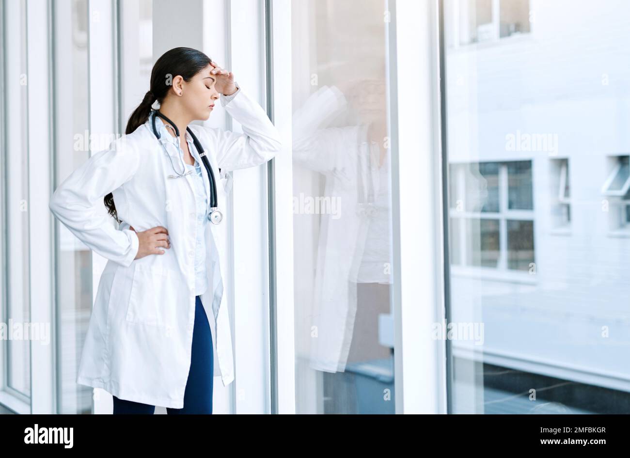 I medici hanno uno dei lavori più stressanti del mondo. una giovane donna che guarda stressato fuori mentre in piedi in una finestra in un ospedale. Foto Stock