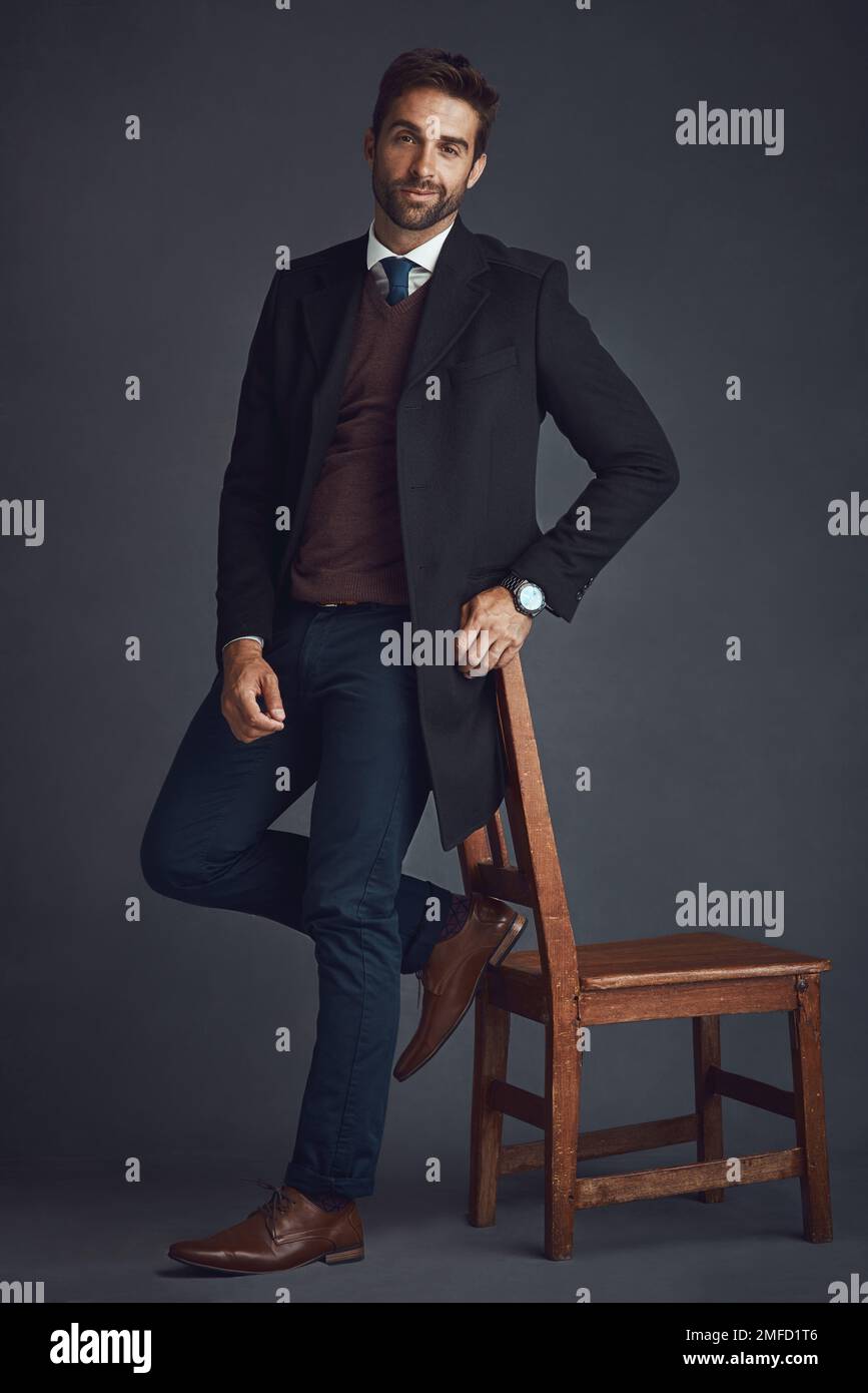I vestiti fanno l'uomo. Studio ritratto di un giovane uomo elegantemente vestito in piedi accanto a una sedia su uno sfondo grigio. Foto Stock