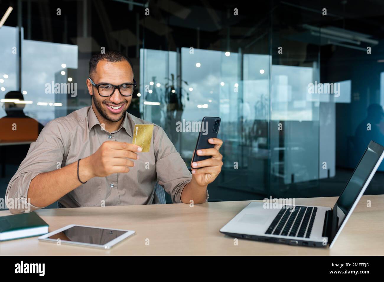 Giovane afro-americano sorridente seduto in ufficio alla scrivania con un computer portatile. È in possesso di una carta di credito, utilizzando il telefono. Foto Stock