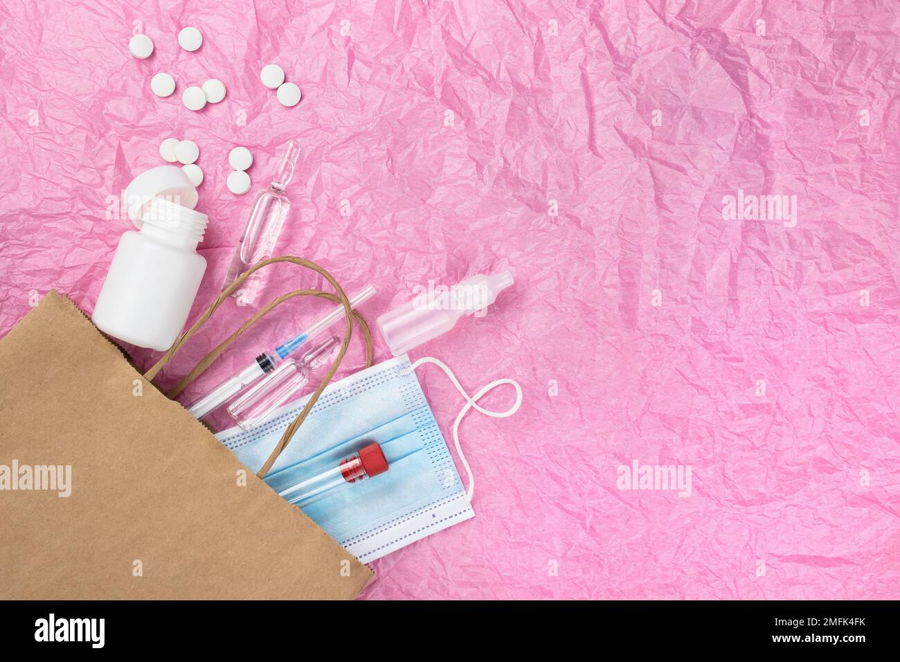 Articoli medici come pillole ampolle, una siringa e una maschera peek fuori da un sacchetto di carta contro uno sfondo di carta rosa stropicciata. Un sacco di spazio vuoto fo Foto Stock